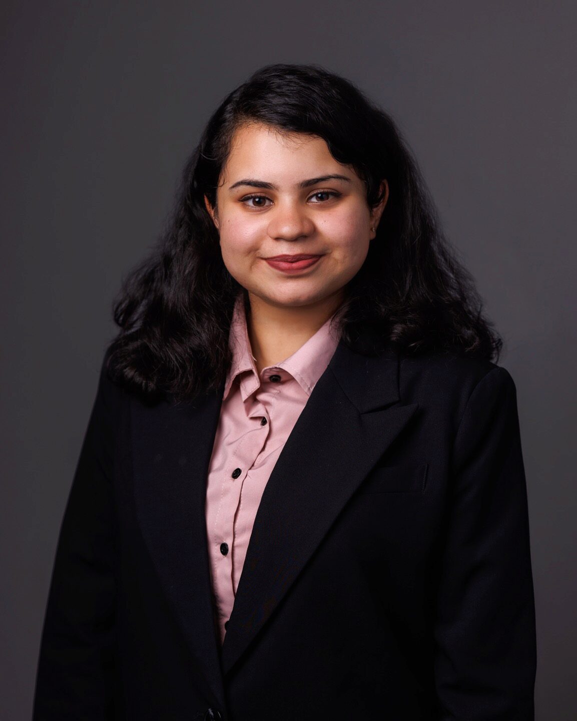 A person posing for a professional headshot, wearing a dark blazer and a pink shirt against a plain background.