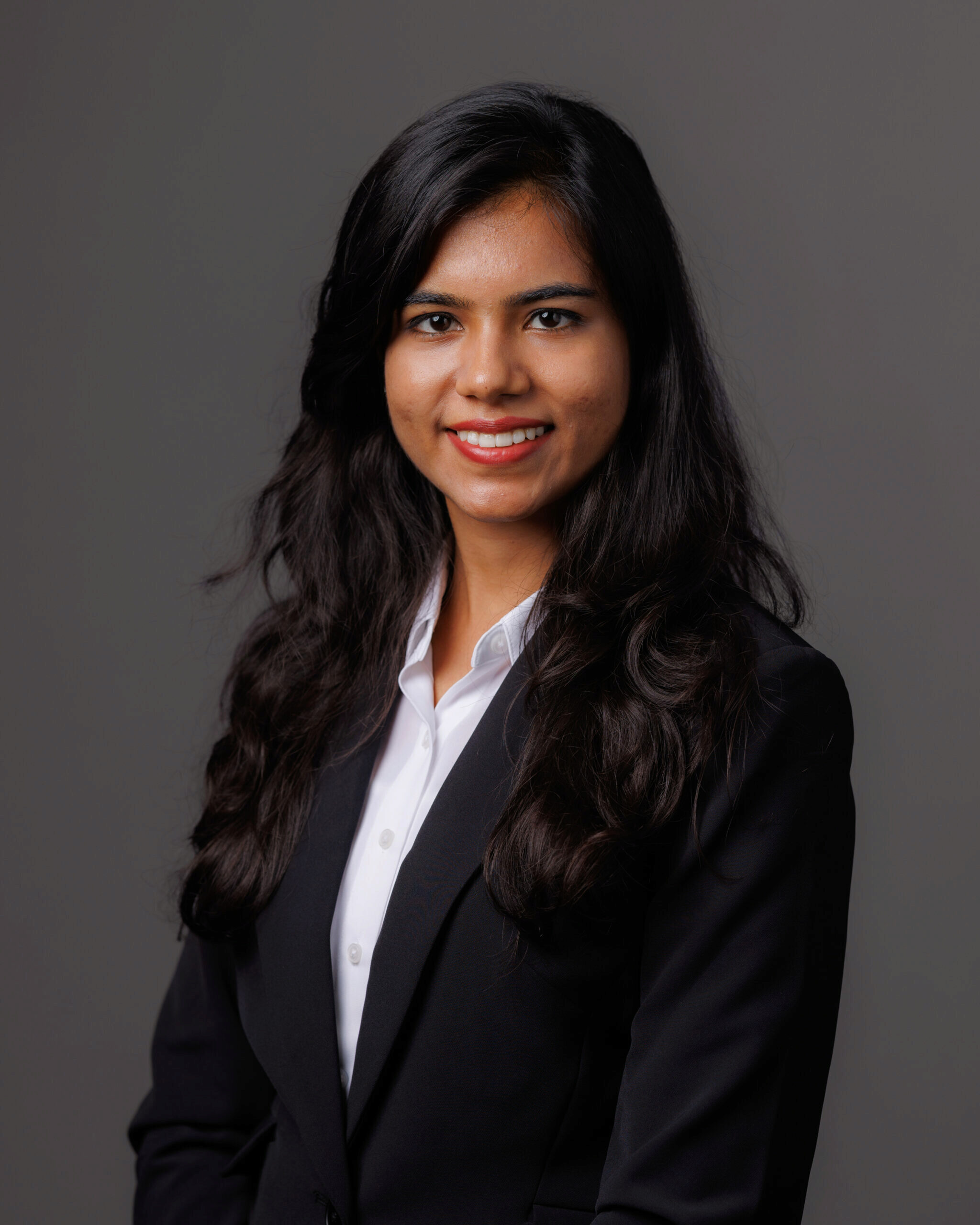 A professional headshot of a person with long dark hair wearing a white shirt and dark blazer, smiling against a plain background.