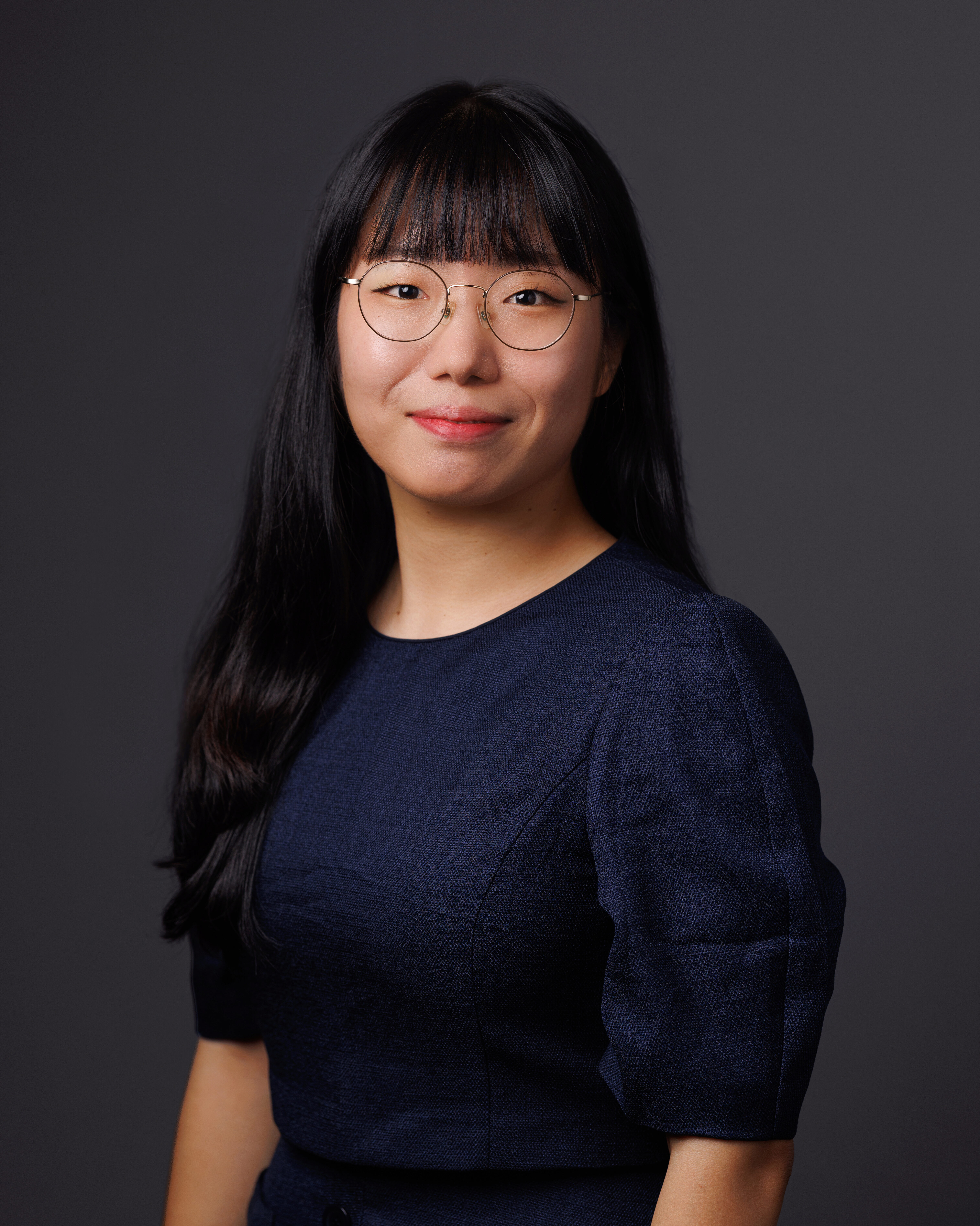 A headshot of a person with long dark hair and glasses, wearing a dark blue top, against a gray background.