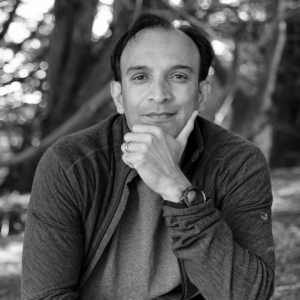 A grayscale headshot of a person outdoors, with a thoughtful expression, resting their chin on their hand. The background is softly blurred.