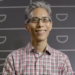 A headshot of a person with glasses, smiling, wearing a plaid shirt. The background features a pattern resembling file drawers.