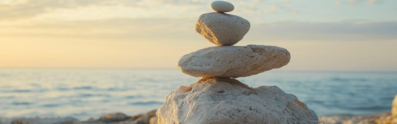 Stacked stones on a beach with a calm ocean and sunset in the background, symbolizing balance and tranquility.