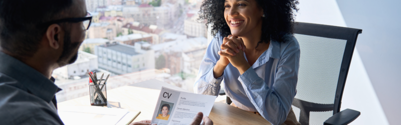 A person in an office chair smiling during a job interview, with another person holding a CV. The setting is an office with a cityscape view from the window.