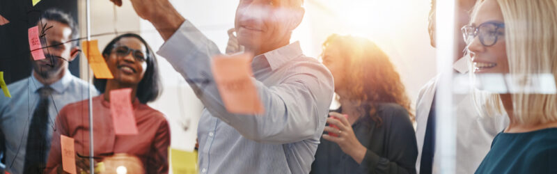 A group of people engaged in a collaborative office meeting, gathered around a glass board with colorful sticky notes. One person is pointing while others look on, smiling. The scene suggests teamwork and brainstorming.